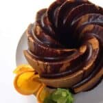 A chocolate-glazed orange poppy seed cake sits on a white plate, garnished with orange slices and green leaves.