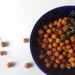 A blue bowl filled with spicy garlic roasted chickpeas garnished with fresh herbs, with a few chickpeas scattered on a white surface nearby.