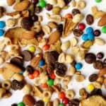 A mix of dried fruit, nuts, spicy trail mix, colorful candy-coated chocolates, chocolate chips, peanuts, and cereal pieces scattered on a white surface.