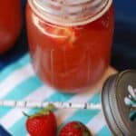 A mason jar filled with refreshing Strawberry Lemonade Shandies sits on a blue-and-white napkin next to a metal lid, two fresh strawberries, and a striped paper straw.