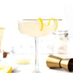 A coupe glass filled with a pale yellow cocktail, garnished with a curled lemon peel on the rim. Bar tools, lemon, and ingredients hint at creative French 75 variations in the background.