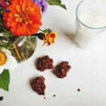 A glass of milk, three No-Bake Nutella Oatmeal Cookies, and a vase with assorted flowers on a white surface.