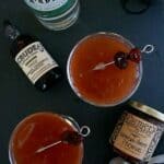 Two Improved Gin + Jam Cocktails, garnished with cherries, sit on a dark surface surrounded by sparkling water, cocktail bitters, and a jar of mountain cherry preserves.
