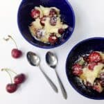 Two blue bowls with cherry clafoutis dusted in powdered sugar, two silver spoons, and three fresh cherries on a white surface.