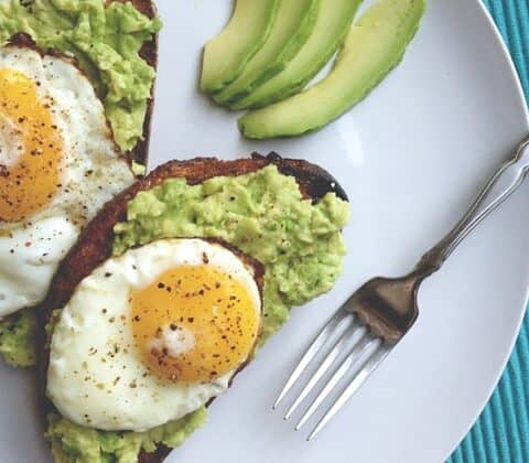 Two slices of avocado toast topped with fried egg and mashed avocado on a white plate, served with fresh avocado slices and a fork on the side.