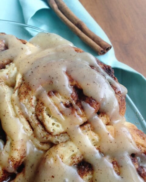 Close-up of Brown Butter Glazed Cinnamon Rolls topped with icing, with two cinnamon sticks and a light blue napkin in the background.