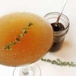 A Ginger Fig Sparkler cocktail, garnished with a sprig of thyme, sits next to a jar filled with a dark mixture and a spoon, while another thyme sprig lies on the table.