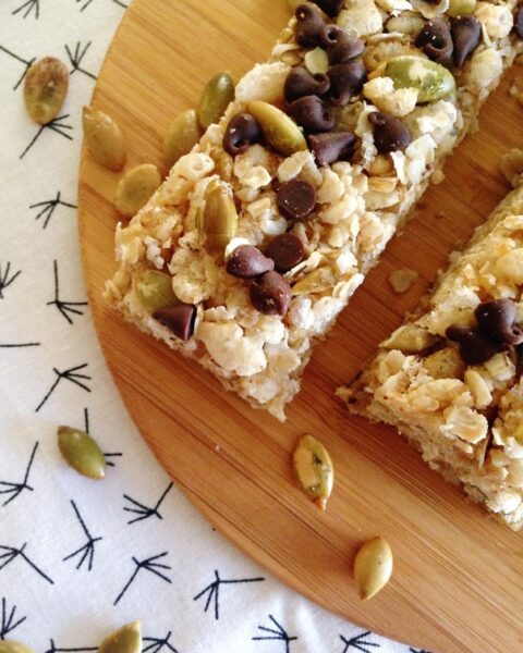 Two Pumpkin Chocolate Chip Granola Bars with oats, pumpkin seeds, and chocolate chips rest on a round wooden board, with seeds scattered on a patterned white cloth.