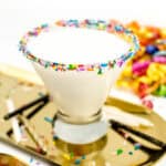 A white cocktail in a stemless martini glass on a gold tray with sprinkles and birthday candles.