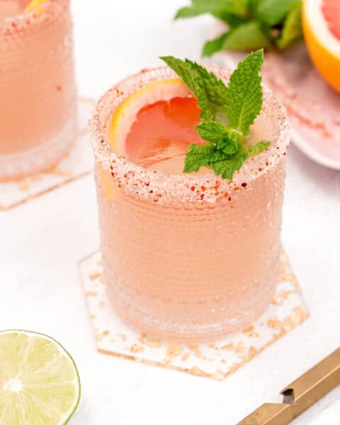 Two glasses of pink Paloma cocktails with salted rims, garnished with grapefruit slices and mint leaves, on a white surface with a lime slice nearby.