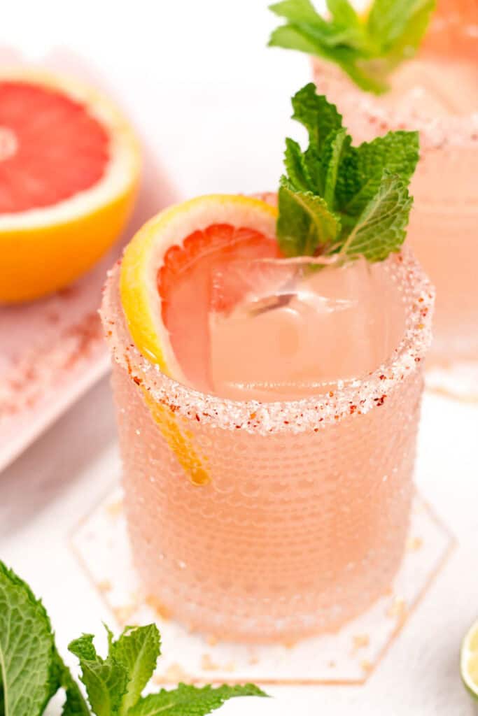 A refreshing Paloma cocktail in a glass rimmed with pink salt, garnished with a mint sprig and a slice of grapefruit.