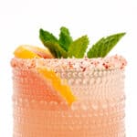 Close-up of a Paloma cocktail with a salted rim, garnished with a lemon wedge and mint leaves, against a white background.