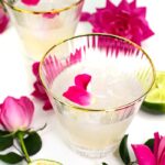 elderflower rose gimlet in a gold-rimmed stemless cocktail glass with pink garden roses and limes on a white background