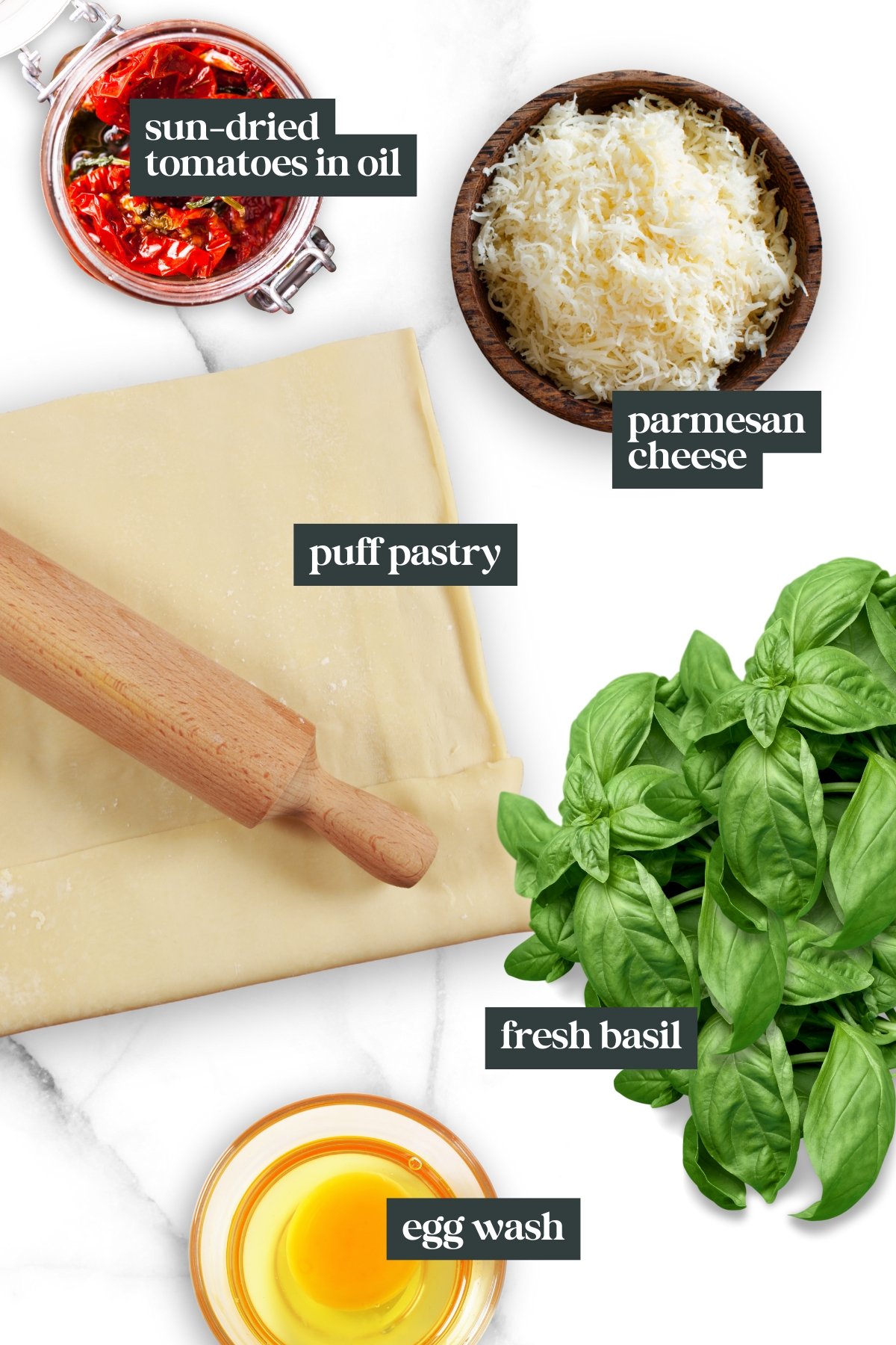 Ingredients for Puff Pastry Pinwheels displayed: sun-dried tomatoes in oil, grated parmesan cheese, puff pastry with rolling pin, fresh basil leaves, and a bowl of egg wash.