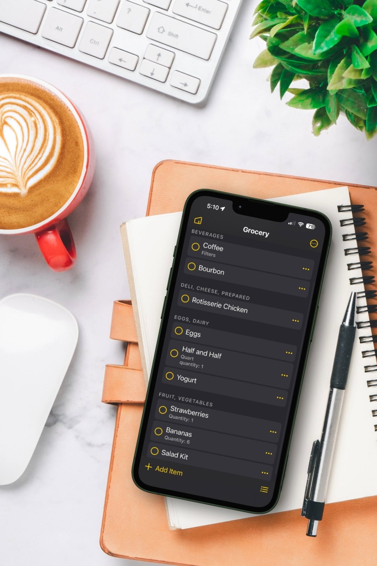 A smartphone displaying one of the best Cooking Apps for grocery lists rests on a notebook next to a pen, a cup of coffee, a plant, and a computer keyboard.