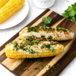 Two ears of grilled Mexican corn, or elote, rest on a wooden board, topped with cheese and herbs. A bowl of cheese, plate of corn, and sprigs of cilantro complement the setting on a light surface.