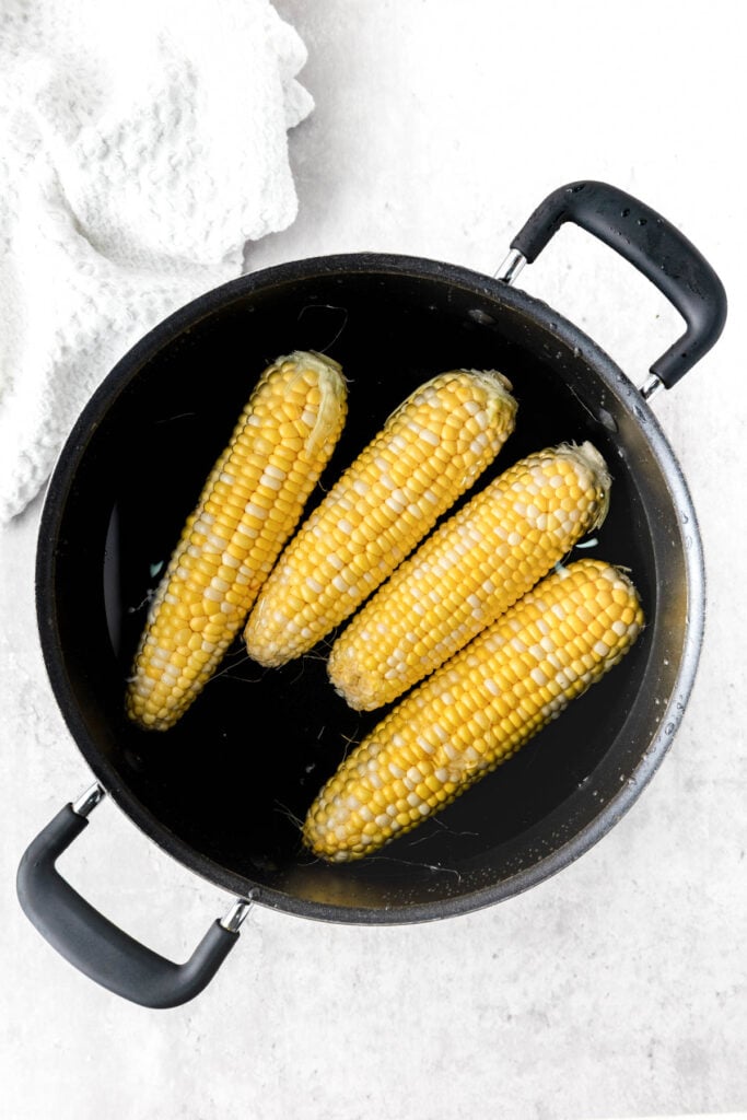 Four ears of corn, perfect for transforming into delicious grilled Mexican corn, are submerged in water inside a black pot with two handles on a light surface, beside a white textured cloth.