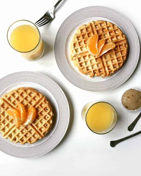 Two plates of Orange Waffles with Vanilla Bean Butter, topped with orange slices, are served alongside two glasses of orange juice, cutlery, and a small round pastry on a white surface.