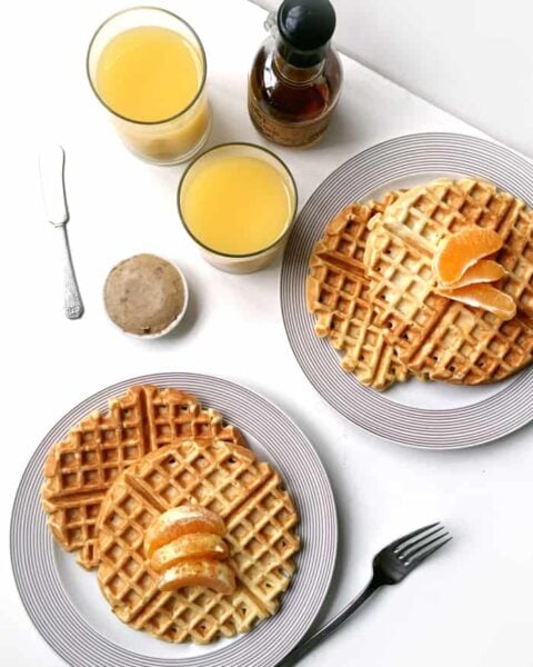 Two plates of Orange Waffles with Vanilla Bean Butter and orange slices, two glasses of orange juice, a bottle of syrup, a butter knife, and a small container of butter sit on a white surface.