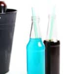 Two glass bottles with straws, featuring bottled tailgate cocktails—one with blue liquid and the other with dark brown—stand beside a black bucket on a white background.