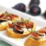 Fig and Goat Cheese Crostini are elegantly arranged on a white tray, featuring slices of bread topped with figs, creamy cheese, and fresh herbs.