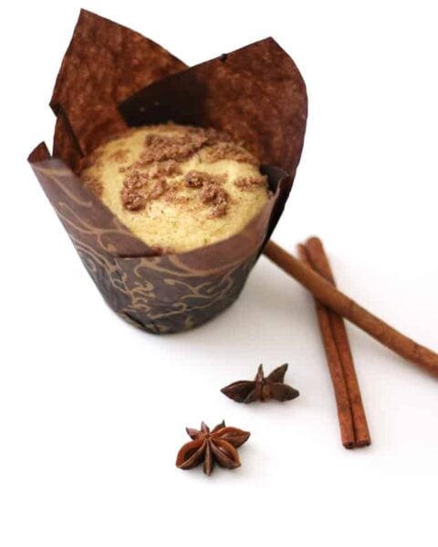 A delightful chai muffin in decorative paper, topped with crumble, sits next to cinnamon sticks and star anise on a pristine white background.