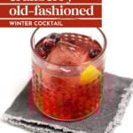 A glass of cranberry old-fashioned cocktail with ice, cranberries, and a lemon peel sits on a gray napkin; text overlay reads "cranberry old-fashioned winter cocktail." Perfect for cozy evenings.