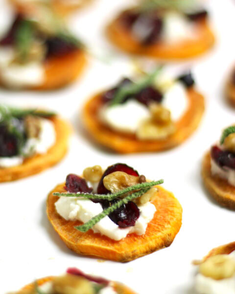 Holiday Sweet Potato Toast Bites feature sweet potato rounds topped with creamy goat cheese, cranberries, walnuts, and fresh herbs on a white surface.