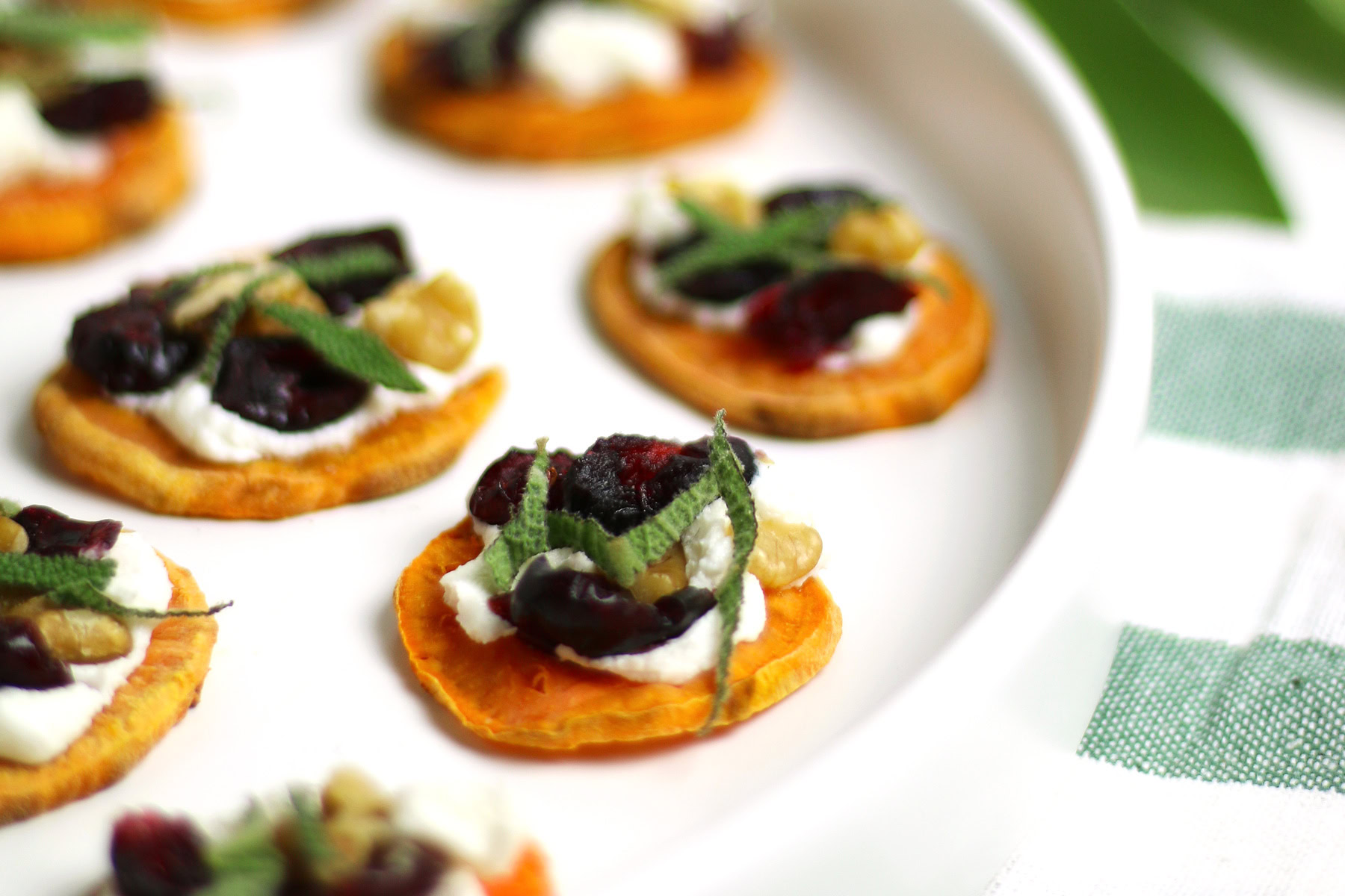 Holiday Sweet Potato Toast Bites: Sweet potato rounds topped with cheese, cherries, walnuts, and mint on a white plate.