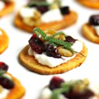 Holiday Sweet Potato Toast Bites: Sweet potato round topped with goat cheese, cranberries, walnuts, and herbs, on a white background.