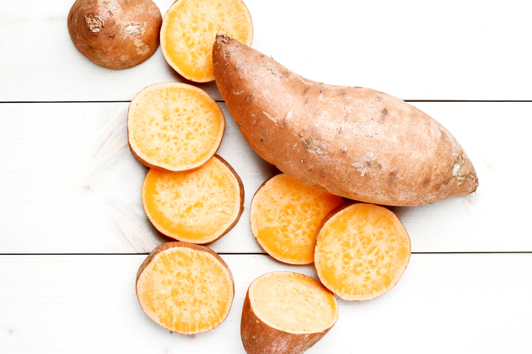 Whole and sliced sweet potatoes on a white wooden surface, perfect for making Holiday Sweet Potato Toast Bites.