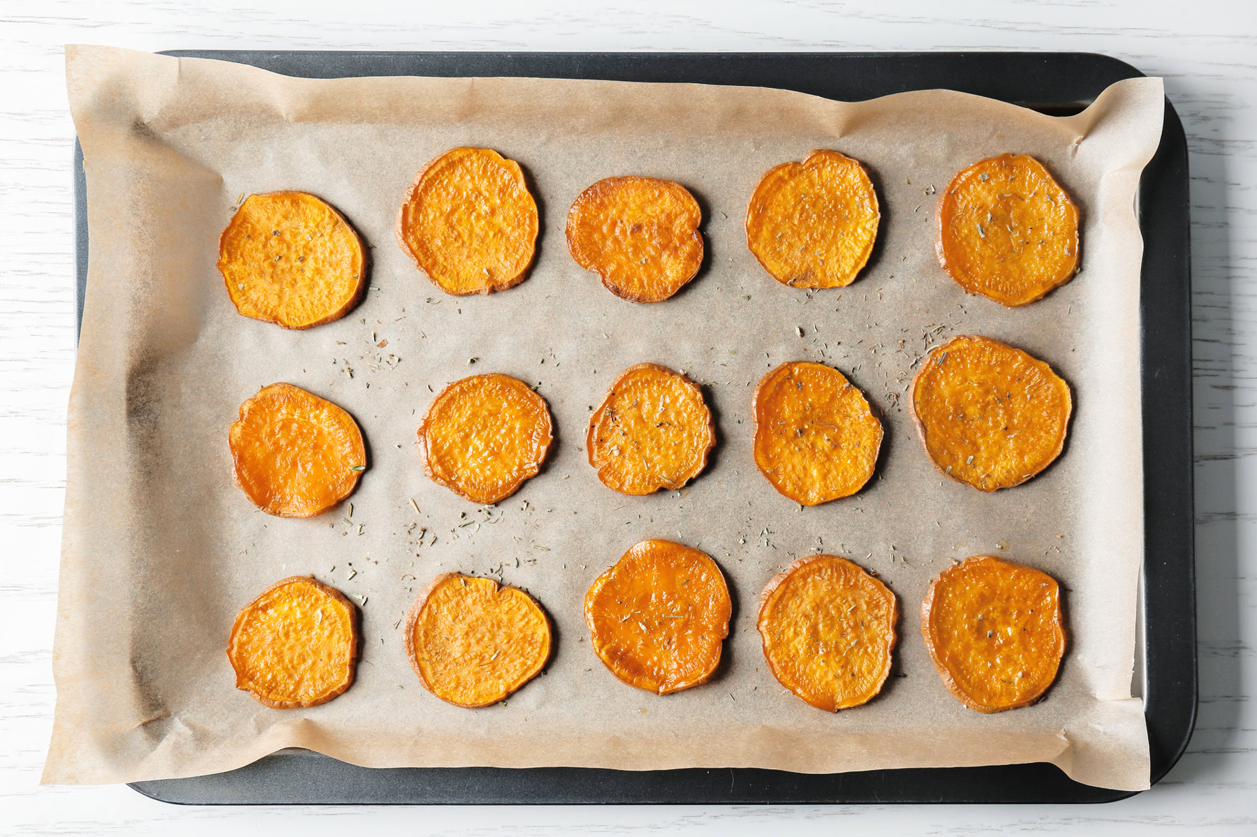 Sixteen baked sweet potato slices on parchment paper, arranged in rows on a black baking tray—these Holiday Sweet Potato Toast Bites make for a festive and delicious appetizer or snack.