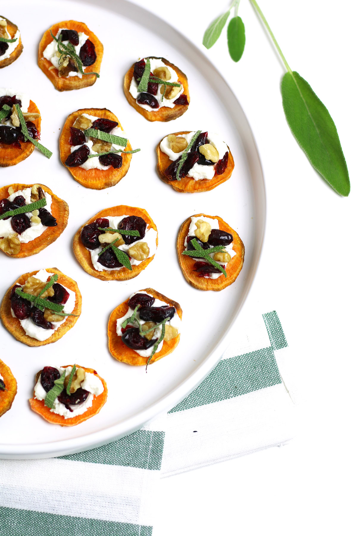 Holiday Sweet Potato Toast Bites: Round sweet potato bites topped with cream, herbs, nuts, and cranberries arranged on a white plate, napkin nearby.