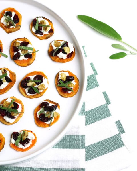 Holiday Sweet Potato Toast Bites are served as round sweet potato bites with toppings on a white plate, set next to a green-striped napkin and fresh sage leaves.