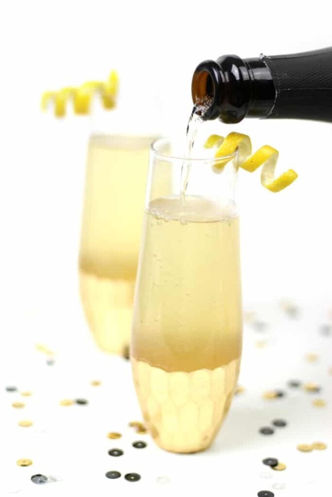 A bottle pours champagne into a flute glass with a lemon twist garnish, crafting an elegant elderflower French 75; another garnished glass and scattered confetti appear in the background.