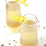 Two champagne flutes filled with a light yellow elderflower French 75, each garnished with lemon twists, sit on a white surface dotted with scattered confetti.