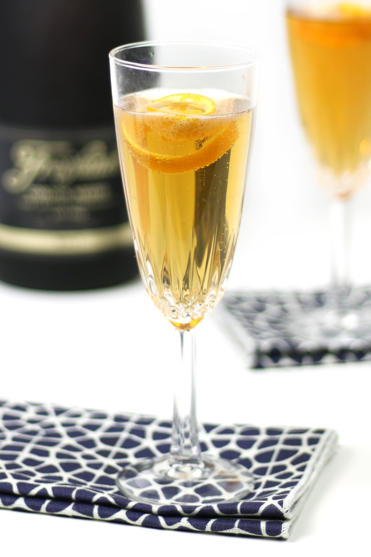 A glass of Seelbach Bourbon Champagne Cocktail with a lemon twist garnish rests on a patterned napkin, while a blurred bottle and another glass set the scene in the background.