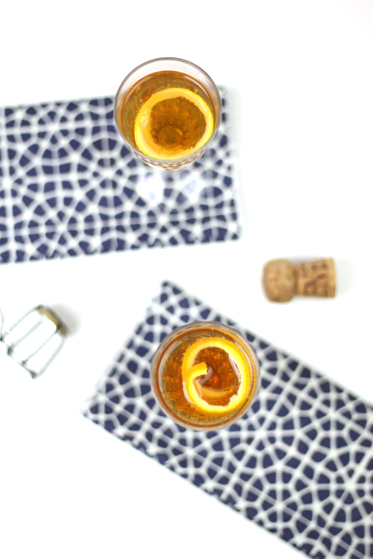 Two glasses of a golden Seelbach Bourbon Champagne Cocktail with orange slices rest on patterned blue and white napkins, next to a cork and wire cage on a white surface.