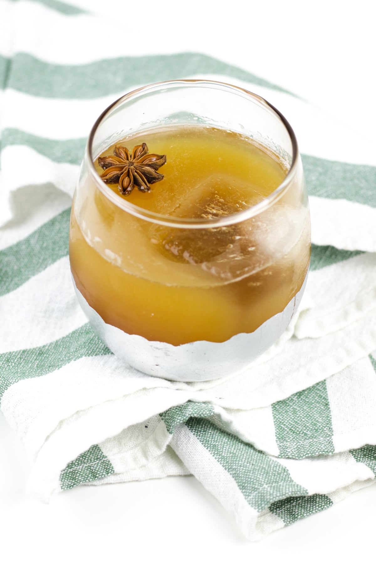 A glass of brown apple cider punch garnished with a star anise pod rests on a green and white striped cloth.