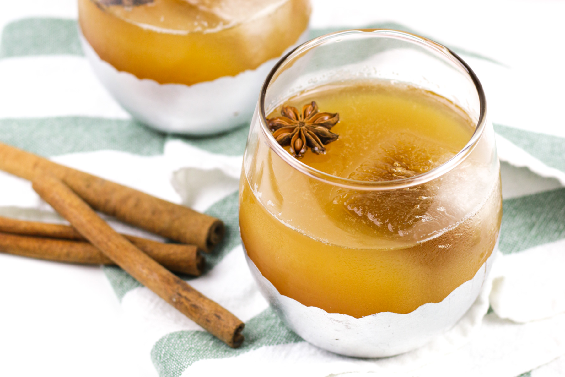 A glass of brown iced apple cider punch garnished with star anise sits beside cinnamon sticks on a white and green striped cloth.