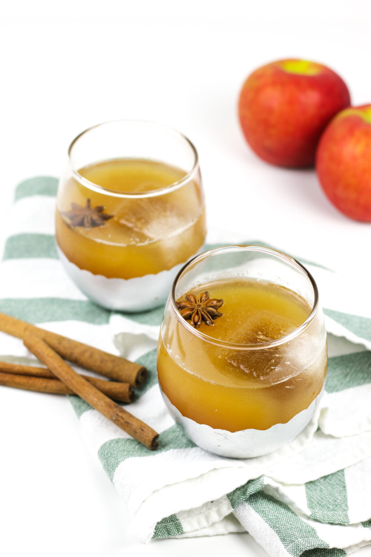 Two glasses of apple cider punch garnished with star anise sit on a striped cloth, with cinnamon sticks and whole apples in the background.