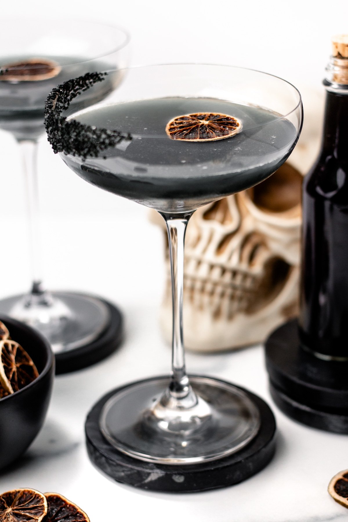 A black cocktail in a coupe glass with a black sugar rim and a dried citrus slice, inspired by apple cider punch, set in front of a skull decoration and a dark bottle.