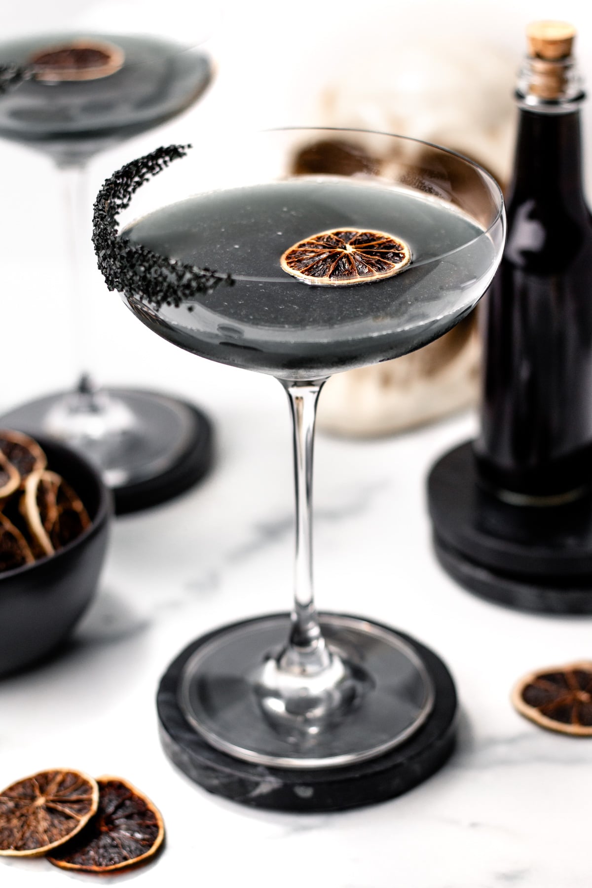 A coupe glass filled with a dark apple cider punch garnished with a dried citrus slice and a rim of black salt, surrounded by dried citrus and a bottle on a marble surface.