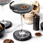 A black cocktail in a coupe glass with a dried citrus slice, black sugar rim, and Halloween-themed decorations—including a skull and bottle—sits on a white surface, perfect for those seeking an eerie twist on classic apple cider punch.