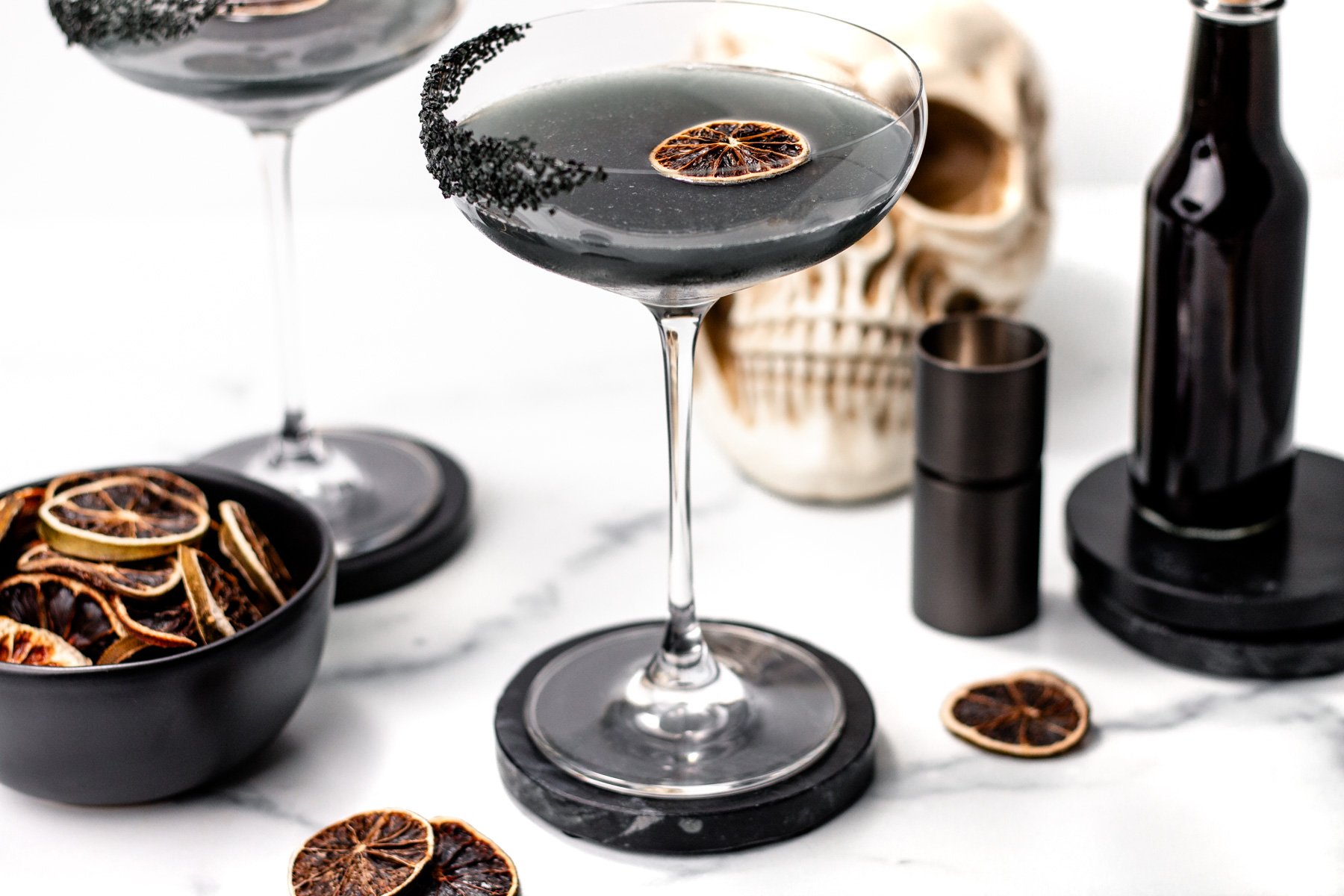 A black cocktail in a coupe glass with a dried citrus slice, black sugar rim, and Halloween-themed decorations—including a skull and bottle—sits on a white surface, perfect for those seeking an eerie twist on classic apple cider punch.