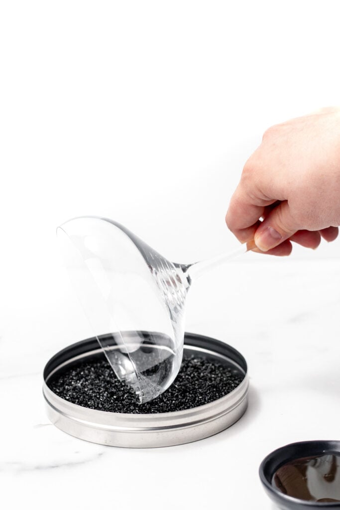 A hand tilts a martini glass, dipping the rim into a shallow tin of black grains—an elegant touch, perhaps preparing it for a festive apple cider punch.