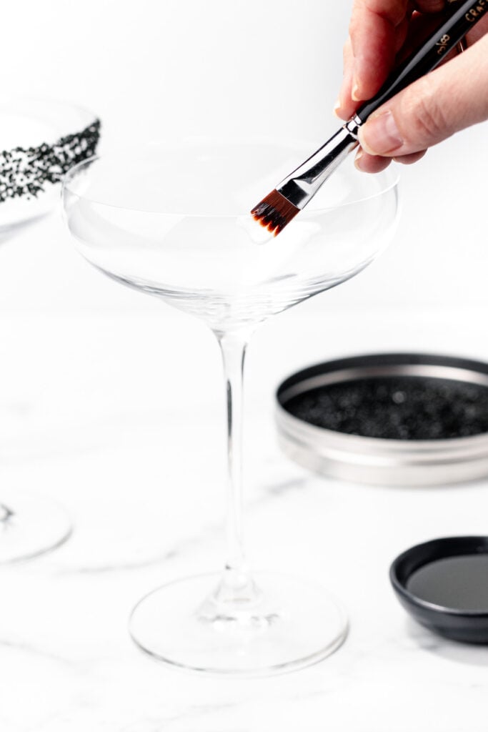 A hand uses a brush to apply liquid for an apple cider punch to the rim of an empty coupe glass, with a dish of black liquid and a tin of black sprinkles nearby.