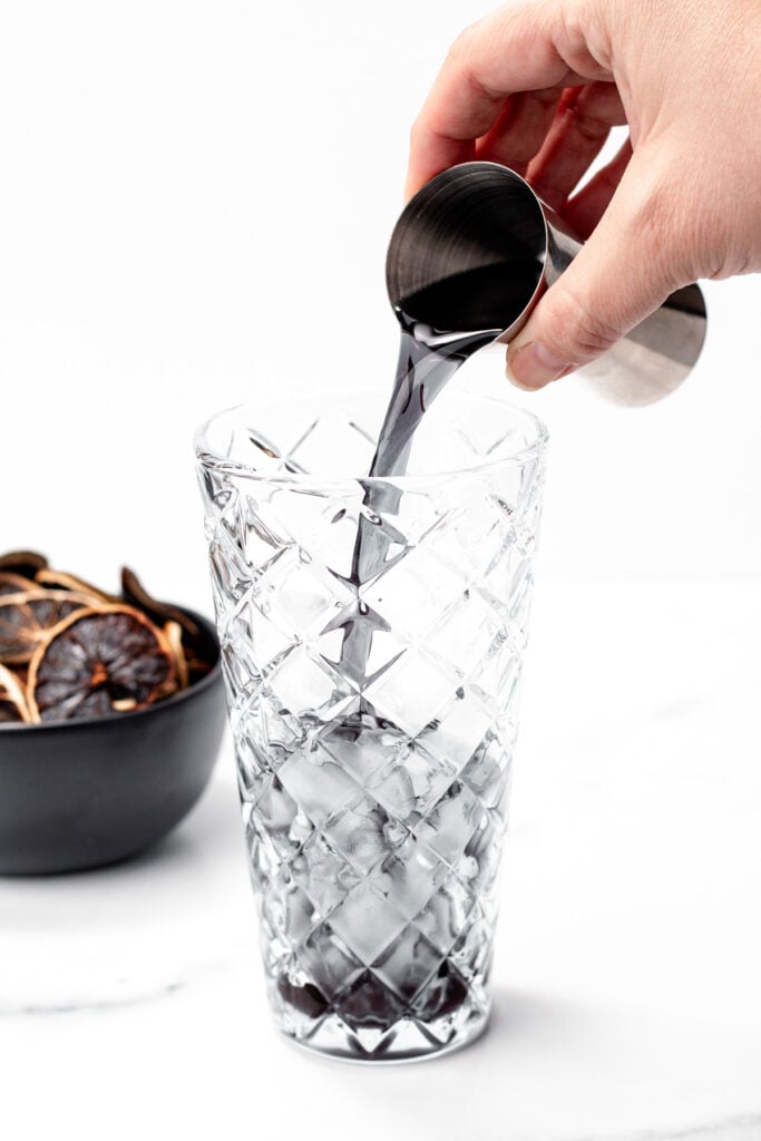 A hand pours a dark liquid from a metal jigger into a tall, textured glass with ice—perfect for serving refreshing apple cider punch. A bowl of dried citrus slices sits in the background.