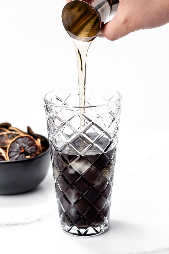 A hand pours a clear liquid from a metal jigger into a tall glass with apple cider punch and ice. A bowl of dried citrus slices sits in the background.