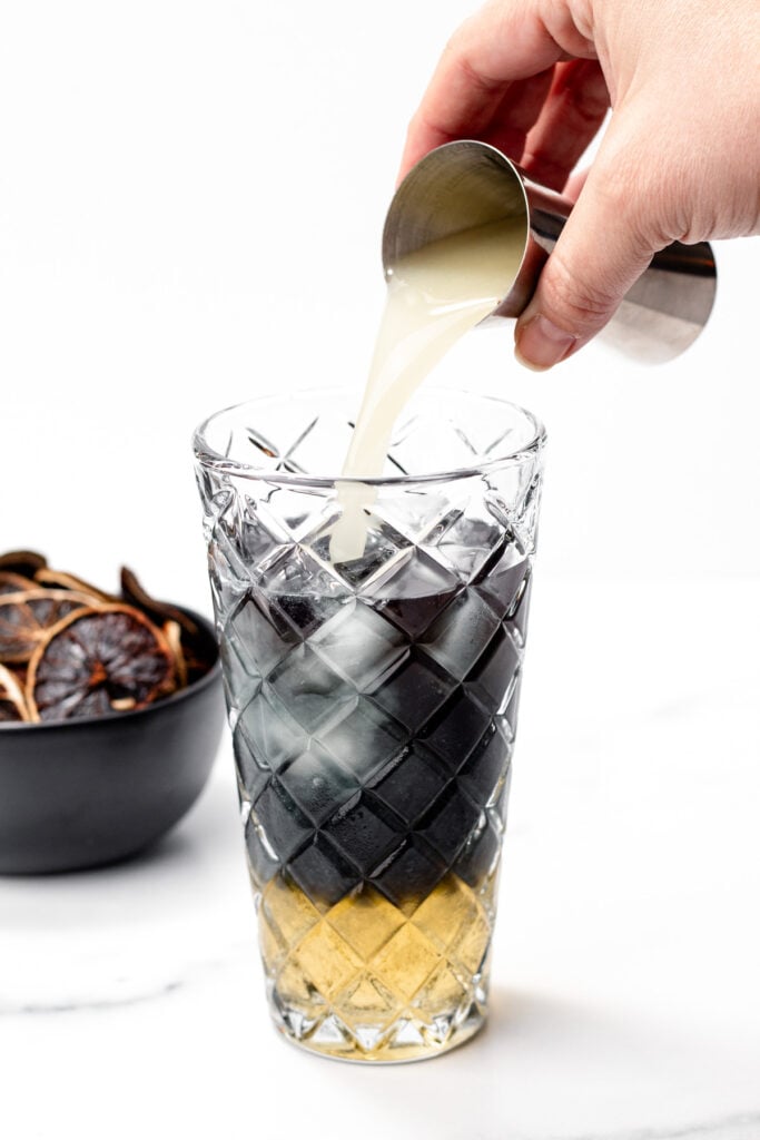 A hand pours a light-colored liquid from a metal jigger into a tall glass of apple cider punch, layered with dark and yellow liquids, while a bowl of dried citrus slices sits in the background.